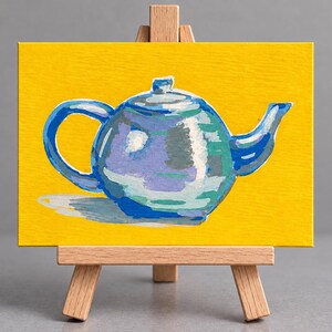 Könnte beinhalten: Ein kleines rechteckiges Leinwandgemälde mit einer blauen Teekanne mit weißen und hellblauen Highlights vor einem einfarbigen gelben Hintergrund. Die Leinwand steht auf einer hölzernen Miniatur-Staffelei mit drei Beinen und einer Querstrebe.