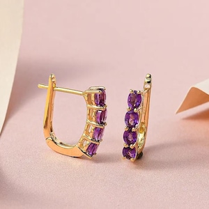 May include: A pair of gold hoop earrings with a row of purple amethyst gemstones. The earrings have a hinged closure.