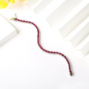 Könnte beinhalten: Ein silbernes Armband mit oval geschliffenen, tiefroten Edelsteinen. Die Edelsteine sind eng aneinandergereiht und bilden eine durchgehende Linie. Das Armband liegt auf einer weißen Oberfläche mit geometrischem Hintergrund.