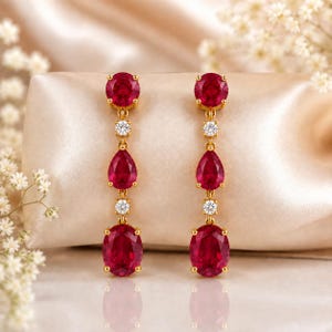 May include: A pair of gold drop earrings featuring a vertical arrangement of a round ruby, a small round diamond, a pear-shaped ruby, another small round diamond, and a large oval ruby, all set in polished yellow gold metal settings.