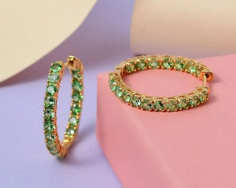 May include: A pair of gold-colored hoop earrings, each encrusted with a row of small, light green gemstones. The earrings are displayed on a pink and purple surface, showcasing their design and color.