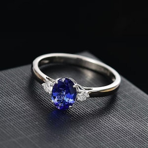 May include: A silver ring with a large oval, deep blue gemstone as the centerpiece. Two smaller, clear gemstones flank the main stone. The ring is set against a dark, textured surface, with a black background.