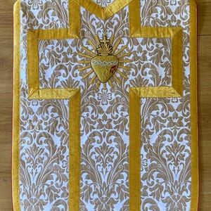 Könnte beinhalten: Ein liturgisches Gewand, wahrscheinlich eine Kasel, mit einem weißen und goldenen Damastmuster. Goldbesatz und ein goldgesticktes Herz-Jesu-Emblem zieren die Vorderseite. Die Kanten sind rot eingefasst.