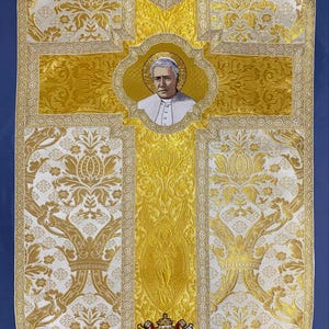 May include: A white and gold liturgical vestment with a cross design. The cross features a portrait of a man in a white robe. The vestment also includes a coat of arms with a lion and an anchor.