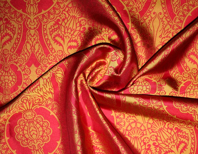 K&ouml;nnte beinhalten: Roter Stoff mit goldfarbenem Paisley-Muster. Der Stoff ist gefaltet und drapiert und zeigt die Textur und den Glanz des Materials.