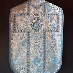 May include: A white and blue liturgical vestment with a floral pattern and a gold embroidered monogram. The vestment has a blue trim and a white lace trim.