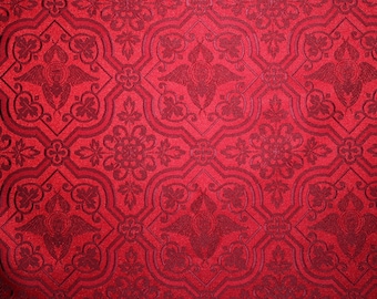 Pode incluir: Tecido vermelho com um padrão repetido de um desenho floral estilizado e um querubim alado. O tecido tem um aspecto texturizado e em relevo.