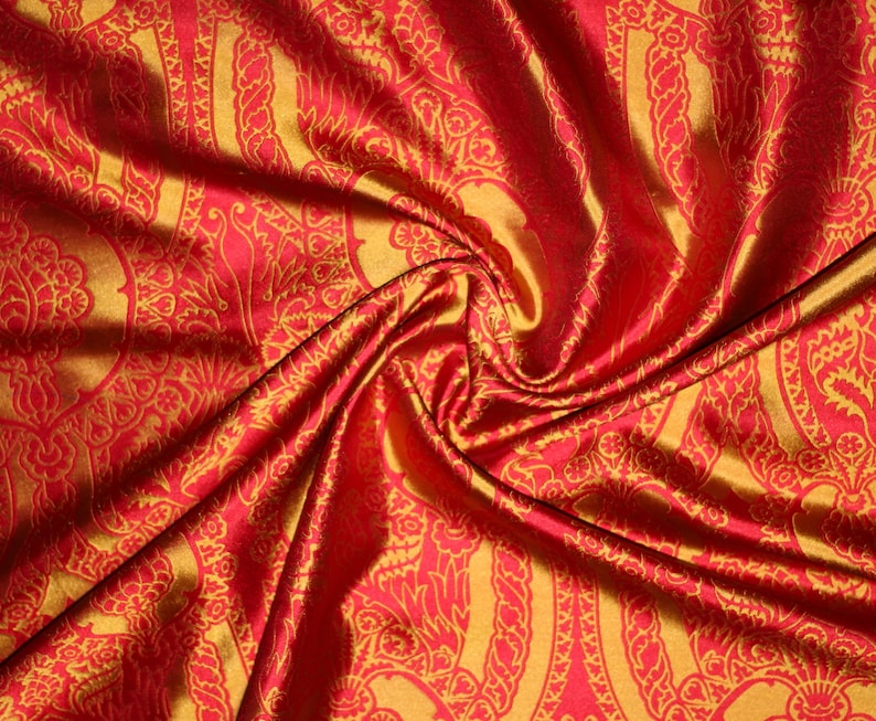 K&ouml;nnte beinhalten: Roter Stoff mit goldfarbenem Paisley-Muster. Der Stoff ist drapiert und gefaltet, wodurch die Textur und der Glanz des Materials sichtbar werden.