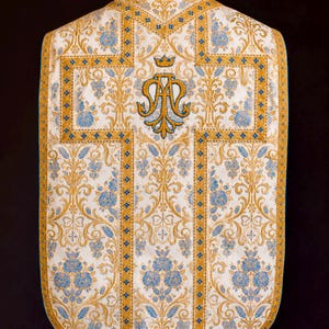 May include: A white liturgical vestment featuring intricate gold and blue floral embroidery. A prominent gold cross design runs vertically and horizontally across the center, topped with a gold crown and the monogram AM in gold and blue thread.