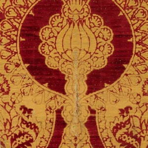 K&ouml;nnte beinhalten: Nahaufnahme eines roten und goldenen Stoffes mit einem sich wiederholenden Muster aus stilisierten Blumen und Bl&auml;ttern. Der Stoff hat eine reiche, strukturierte Optik und Haptik.