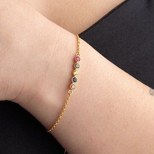 May include: A gold chain bracelet with a row of colorful gemstones. The bracelet has a delicate chain and is adorned with small, round stones in shades of blue, green, pink, and yellow.