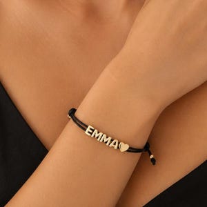 Könnte beinhalten: Ein schwarzes Kordelarmband mit goldfarbenen Metallbuchstabenperlen, die den Namen EMMA bilden, gefolgt von einem kleinen herzförmigen Anhänger in Goldoptik. Das Armband hat verstellbare schwarze Kordelenden mit kleinen goldenen Perlen an den Enden.