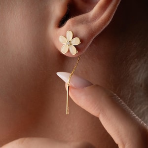 Könnte beinhalten: Nahaufnahme eines blütenförmigen Ohrrings in Gold mit einem kleinen Diamanten in der Mitte. Eine goldene Kette erstreckt sich von dem Ohrring, mit einem goldenen Stab am Ende. Der Ohrring befindet sich im Ohr einer Person. Die Blume hat ein weißes Design.