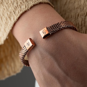 May include: Copper-colored cuff bracelet with a woven chain design. The bracelet has a rectangular clasp with the letter "G" engraved on one end and the letter "M" on the other. The bracelet is worn on a wrist.