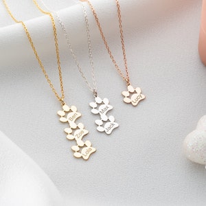 Könnte beinhalten: Gold-, Silber- und Roségold-Halsketten mit Pfotenabdruck-Anhängern. Jeder Anhänger hat eingravierte Namen. Die Halsketten werden auf einer weißen Oberfläche mit einem weichen, drapierten Stoffhintergrund präsentiert. Eine rosa Schmuckschatulle ist im Hintergrund.