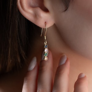 Könnte beinhalten: Goldene Ohrringe mit fünf bunten Edelsteinen. Die Edelsteine sind grün, rosa, blau und gelb.