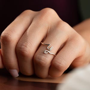 Pode incluir: Um anel de tom prateado com uma banda de fio fino e contínuo que forma uma letra cursiva estilizada. O metal tem um acabamento liso e polido, e o design é aberto, envolvendo o dedo com uma estética delicada e minimalista.