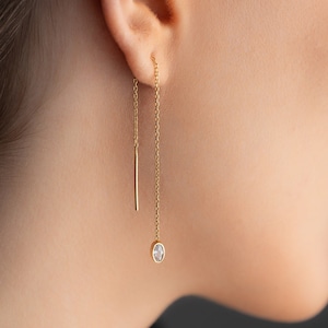 Può includere: Un paio di orecchini a catena d'oro con una piccola pietra preziosa trasparente all'estremità di una catena e una sottile barra d'oro all'estremità dell'altra catena.