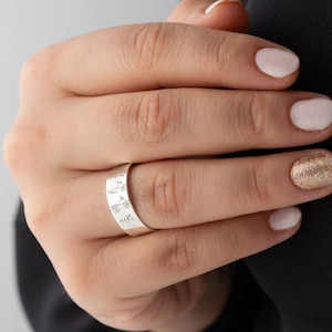 May include: A silver ring with a delicate floral design engraved around the band. The ring is worn on a finger with a light pink nail polish.