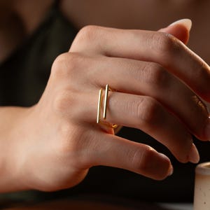 May include: A gold-toned ring featuring an open, geometric design with two parallel vertical bars connected by a horizontal base. The ring has a polished, smooth metallic finish and is worn on a finger, showcasing its clean, linear structure.