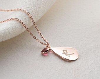 May include: A rose gold necklace with a teardrop pendant featuring a cursive letter "K" and a small pink gemstone.