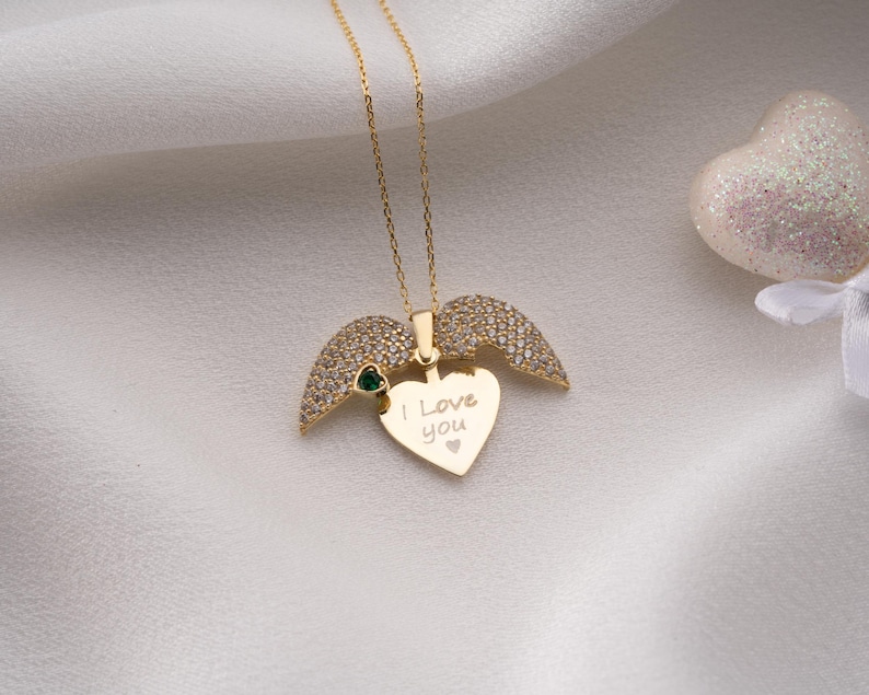 Peut inclure: Un collier dor&eacute; avec un pendentif en forme de c&oelig;ur et des ailes d'ange. Le pendentif porte l'inscription "I Love You". Les ailes sont incrust&eacute;es de petites pierres transparentes. Une petite pierre verte est sertie &agrave; la base des ailes.
