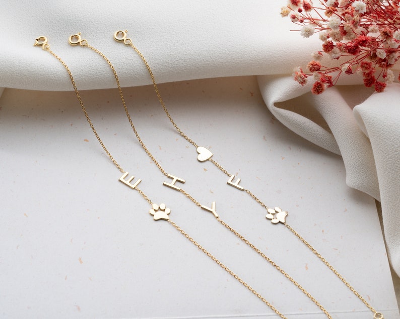 Puede incluir: Tres pulseras de cadena de oro con diferentes dijes. Los dijes incluyen un coraz&oacute;n, una huella de pata y las letras E, H e Y.