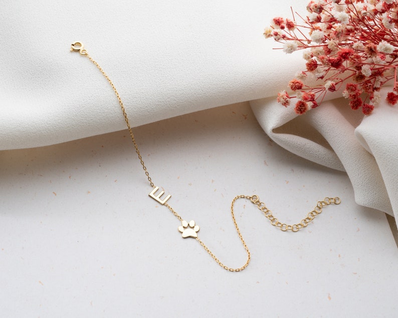 Puede incluir: Un brazalete de cadena dorada con un peque&ntilde;o dije de huella de pata y un dije con la letra 'E'. El brazalete est&aacute; sobre una superficie blanca.