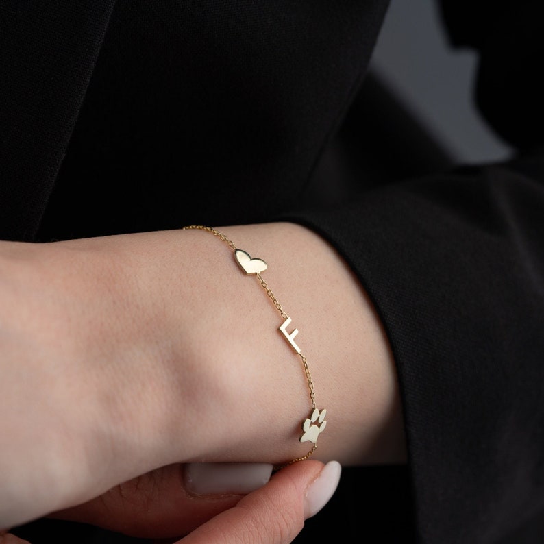 Puede incluir: Un brazalete de cadena dorada con dijes de coraz&oacute;n, letra F y huella de pata.