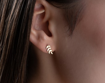 Könnte beinhalten: Ein Paar goldene Blattohrringe. Die Ohrringe sind klein und zart und haben ein schlichtes Design aus einem Zweig mit Blättern.