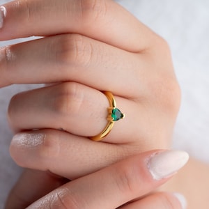 May include: A gold ring with a green heart-shaped gemstone. The ring is on a person's finger.