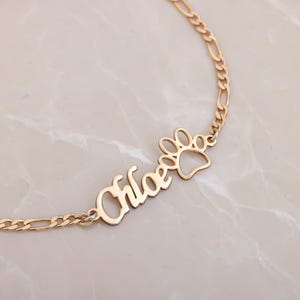 Könnte beinhalten: Goldfarbene Halskette mit dem Namen "Chloe" und einem Pfotenabdruck-Anhänger. Die Halskette hat eine filigrane Kette und wird auf einer Marmoroberfläche präsentiert. Der Pfotenabdruck-Anhänger verleiht dem personalisierten Schmuck eine verspielte Note.