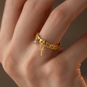 Op de afbeelding: Een goudkleurige ring met een schakelkettingontwerp. De ring heeft een kleine, donkerblauwe edelsteen en een gouden ankh-bedel. De ring wordt om een vinger gedragen en toont de ingewikkelde details en elegante stijl.