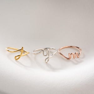 Könnte beinhalten: Drei dünne Metallringe mit Buchstaben in Schreibschrift. Der erste Ring ist goldfarben mit dem Buchstaben J, der zweite silberfarben mit dem Buchstaben y und der dritte roségoldfarben mit dem Buchstaben m, alle aus Draht gefertigt.