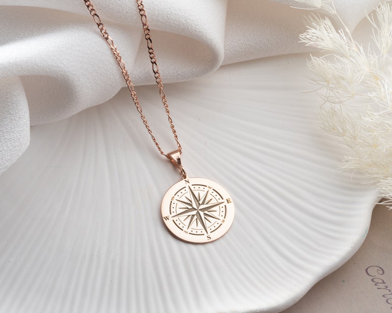 May include: A rose gold necklace with a compass pendant. The compass is round and has a detailed design with the letters N, S, E, and W.