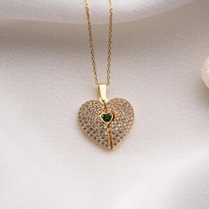 Peut inclure: Un collier dor&eacute; avec un pendentif en forme de c&oelig;ur. Le pendentif est incrust&eacute; de petites pierres transparentes et comporte une petite pierre verte au centre. Le collier est pos&eacute; sur un fond blanc textur&eacute;.