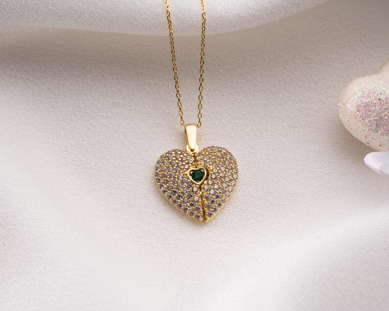 Peut inclure: Un collier dor&eacute; avec un pendentif en forme de c&oelig;ur. Le pendentif est incrust&eacute; de petites pierres transparentes et comporte une petite pierre verte au centre. Le collier est pos&eacute; sur un fond blanc textur&eacute;.