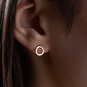 May include: A close-up of a gold-colored, open-circle stud earring. The earring is a simple, minimalist design, and is worn on an ear. The earring is a small, delicate piece of jewelry.