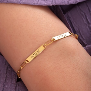 May include: A gold-colored bracelet with rectangular nameplates. The nameplates are linked together with small chain links. The names "Alice" and "Maya" are engraved on the nameplates. The bracelet is worn on a person's wrist.