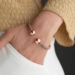 May include: A rose gold cuff bracelet with a woven chain design and two spherical end caps. The open design allows for easy wear. The bracelet is shown on a person's wrist.