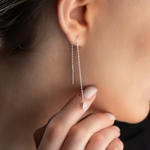 Op de afbeelding: Zilveren kettingoorbellen met een klein, helder edelsteentje aan het einde van de ketting. De oorbellen hangen aan de oorlel.