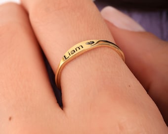 May include: A gold-colored ring with the name "Liam" engraved on it, along with a small heart symbol. The ring is simple and elegant, with a smooth, polished finish. The ring is shown on a finger.