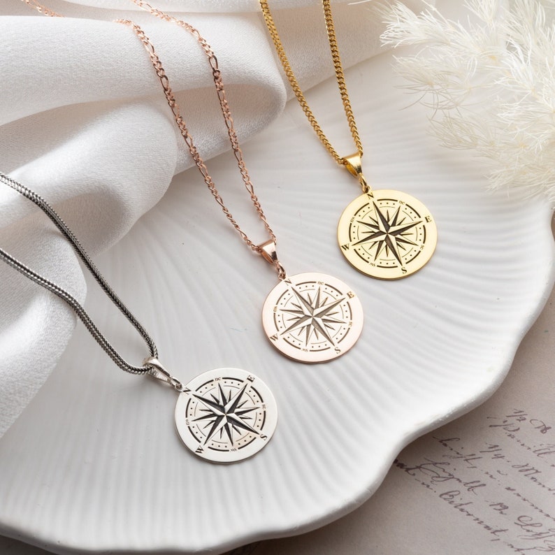 May include: Three silver necklaces with compass pendants. The necklaces are on a white surface with a piece of paper with writing on it. The compasses are in different colors: silver, gold, and rose gold.