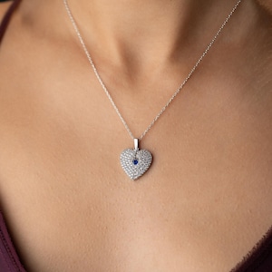 Peut inclure: Un collier en argent avec un pendentif en forme de c&oelig;ur. Le pendentif est incrust&eacute; de petits cristaux transparents et comporte une petite pierre pr&eacute;cieuse bleue ronde au centre. Le collier est port&eacute; sur une poitrine.