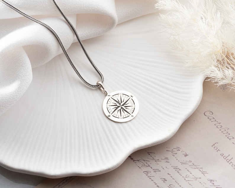May include: A silver necklace with a compass pendant. The compass is a circle with a star in the center and lines radiating out from the star.