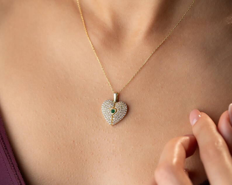 Peut inclure: Un collier dor&eacute; avec un pendentif en forme de c&oelig;ur. Le pendentif est incrust&eacute; de petites pierres claires et d'une pierre verte au centre. Le collier est port&eacute;, avec la cha&icirc;ne visible.
