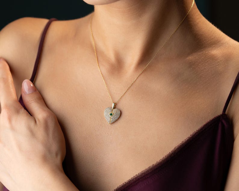 Peut inclure: Un collier dor&eacute; avec un pendentif en forme de c&oelig;ur. Le pendentif est incrust&eacute; de petites pierres claires et comporte une seule pierre verte au centre. Le collier est port&eacute; sur un fond sombre.