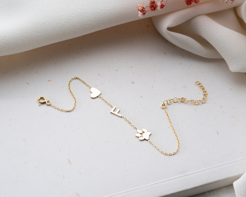 Puede incluir: Un brazalete de cadena dorada con un coraz&oacute;n, una letra F y un dije de huella de pata.