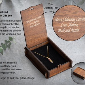 May include: A wooden gift box with a hinged lid. The lid is engraved with the text "Merry Christmas, Caroline Love, Shalena, Mark and Austin". The box is open and a gold necklace with a multi-colored gemstone pendant is inside.