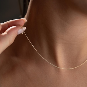 May include: A delicate gold chain necklace. The fine chain is held by a hand, showcasing its subtle design. The necklace is a simple, elegant piece, perfect for everyday wear. The gold color adds a touch of sophistication.